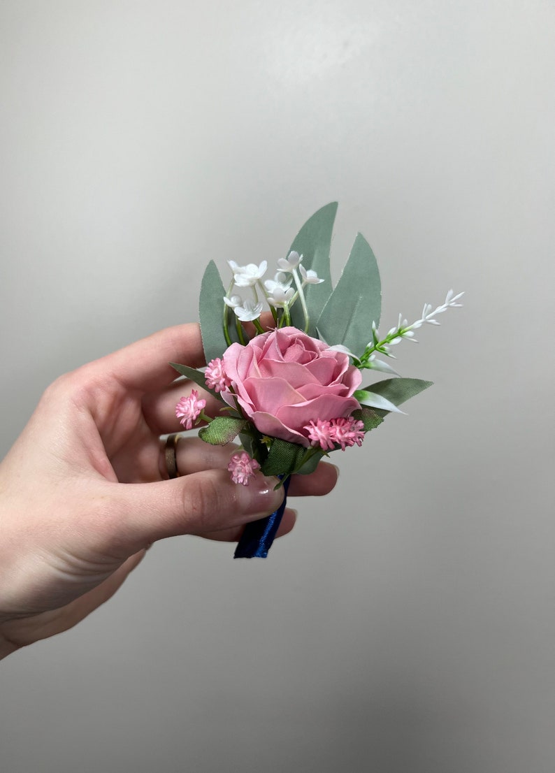 May include: A pink rose boutonniere with white and pink flowers and green leaves. The boutonniere is tied with a blue ribbon.