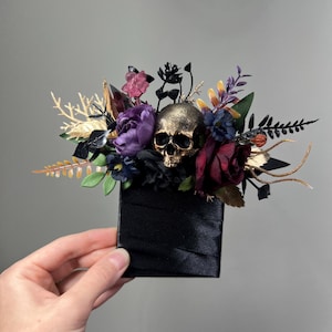 May include: A decorative hair accessory featuring a central metallic gold skull surrounded by an arrangement of artificial flowers in shades of deep purple, burgundy, and black, accented with gold-toned foliage on a black satin-covered base.