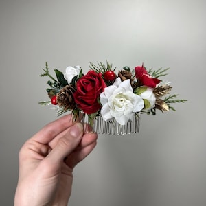 May include: A festive hair comb featuring red and white roses, pinecones, and berries. The floral arrangement is accented with green foliage and gold-colored accents, all attached to a silver comb.