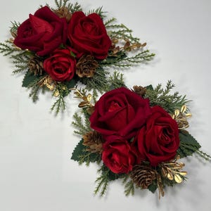 Op de afbeelding: Twee rode rozen bloemstukken met dennentakken en gouden accenten. De rozen zijn in een cluster gerangschikt met dennentakken en gouden accenten.