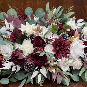 May include: A floral arrangement featuring white, burgundy, and cream-colored flowers, accented with green and gold foliage. The arrangement includes roses, dahlias, and pinecones, creating a festive and elegant display. Perfect for weddings or home decor.