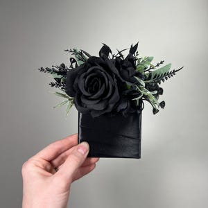 May include: A single black rose with green foliage in a black satin box.