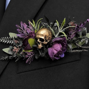 May include: A boutonniere featuring a metallic gold-toned skull centrepiece nestled among dark purple fabric flowers, black foliage, and green leaves. The arrangement is pinned to the chest pocket of a solid black suit jacket with a light blue shirt.