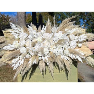 May include: A large, white floral arrangement with pampas grass. The arrangement is made up of white roses, white carnations, and white greenery. The pampas grass is a light brown color and is arranged in a fan shape.