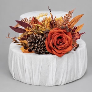 Puede incluir: Una base de pastel blanca de dos niveles decorada con un arreglo floral de temática otoñal. El adorno presenta una gran rosa color óxido, una piña marrón, varias hojas secas en tonos naranja y amarillo, y pequeñas bayas de color rojo oscuro.
