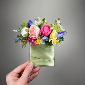 以下が含まれることがあります： 鮮やかな造花のアレンジメントが詰め込まれた、小さな淡い緑色のサテンポーチ。ピンク、白、黄色、青の花と緑の葉が含まれています。ポーチは灰色の背景に対して保持されています。