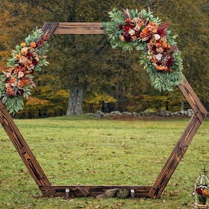 May include: A rustic wooden hexagonal wedding arch decorated with vibrant fall-colored flowers and greenery. The arch is set up on a grassy field, with trees in the background. The arch is approximately 8 feet tall.