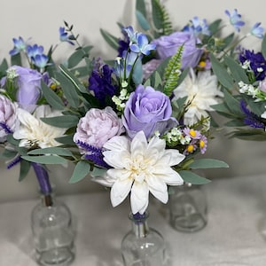 May include: Three bouquets of purple, white and blue artificial flowers in clear glass vases. The flowers include roses, dahlias and lavender.