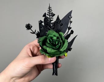 May include: A dark green rose with black foliage and stems. The rose is a paper flower and is part of a boutonniere.