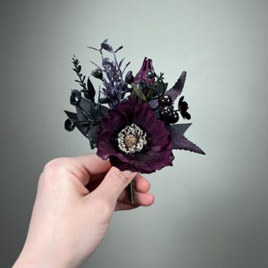 May include: A boutonniere featuring a deep purple poppy with a textured silver center, surrounded by dark purple and black foliage, small black berries, and thin sprigs. The floral arrangement is composed of dark, moody tones and varied textures.