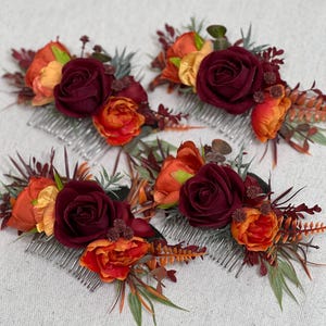 May include: Four floral hair combs with burgundy roses, orange and yellow blossoms, and greenery. The combs have silver-colored teeth and are arranged on a white surface. The flowers are clustered together.