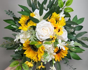 Pode incluir: Um buquê de noiva com girassóis, rosas brancas e folhagem. O buquê é segurado por uma mão.