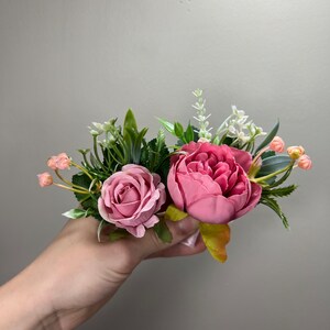 May include: A bouquet of pink and white artificial flowers with green leaves. The flowers are arranged in a hand.