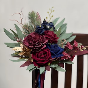 May include: A bouquet of burgundy and navy blue roses, peonies, and other flowers with green and gold foliage. The bouquet is tied with burgundy and navy blue ribbons and rests on a dark wooden chair.