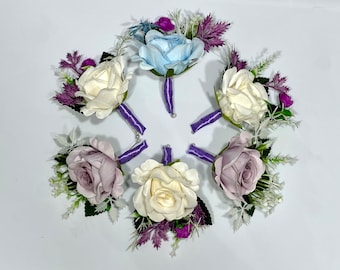 Op de afbeelding: Zes kunstbloemen boutonnières met paarse, witte en lichtblauwe rozen. Elke boutonnière heeft een paars lint en is versierd met kleine paarse bloemen en groen.