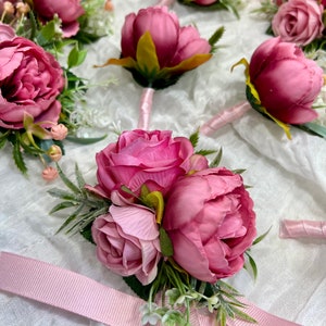 May include: A set of pink silk flower boutonnieres with green leaves and white accents. The boutonnieres are tied with pink ribbon.