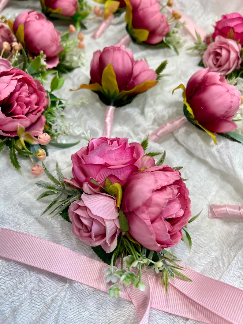 May include: A set of pink silk flower boutonnieres with green leaves and white accents. The boutonnieres are tied with pink ribbon.