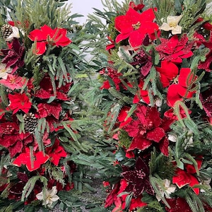 May include: Close-up of two Christmas wreaths with vibrant red poinsettias, pine cones, and evergreen foliage. The wreaths feature a mix of textures and shades of red, creating a festive holiday display.