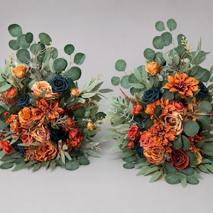 May include: Two floral arrangements featuring a mix of burnt orange and deep navy blue roses and dahlias, accented with various green eucalyptus leaves and slender foliage. The bouquets are composed of artificial flowers in warm autumn tones.