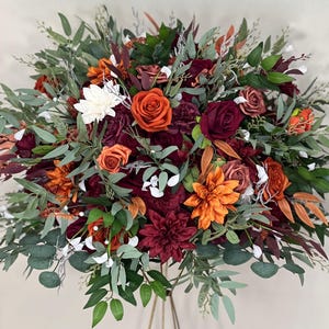 May include: A large floral arrangement with orange, burgundy, and white flowers, complemented by green foliage. The arrangement includes roses, dahlias, and other blooms, creating a full display. The flowers are arranged on a gold metal stand, creating a striking visual.