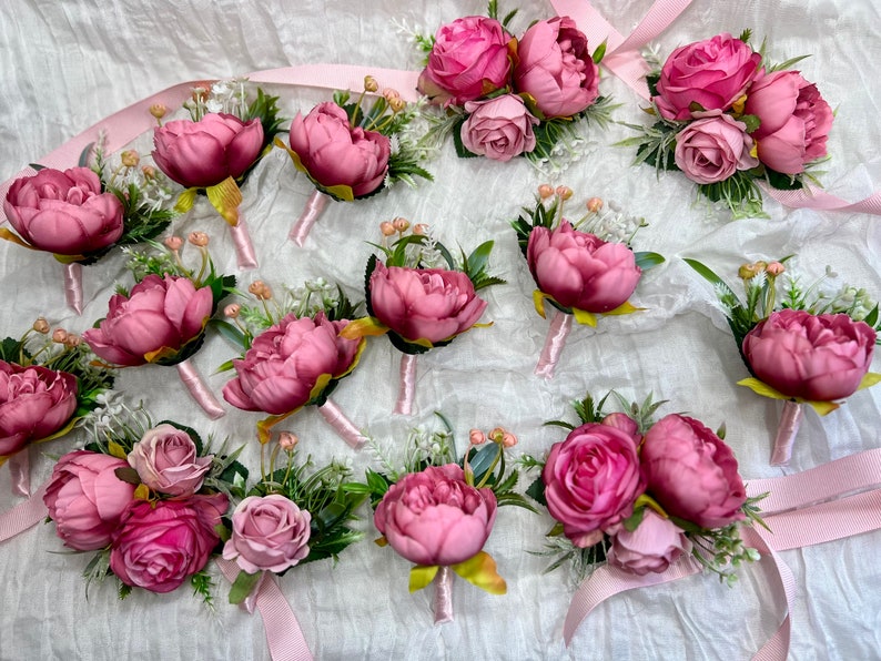 May include: A collection of 15 pink and green floral boutonnieres with a satin ribbon. The boutonnieres are made with silk flowers and greenery.