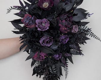 May include: A dark, gothic-style bouquet featuring black and deep purple flowers and foliage. The arrangement includes roses, peonies, and other blooms, with accents of fern-like leaves. The bouquet is held against a plain white background.