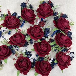 May include: A collection of burgundy rose boutonnieres with blue and white accents. Each boutonniere features a large, velvety rose, surrounded by small blue flowers and greenery. The stems are wrapped in blue ribbon, creating a classic floral arrangement.