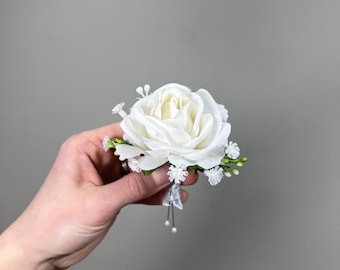 Pode incluir: Um botão de rosa branca com folhas verdes e detalhes brancos. O botão é segurado na mão.