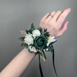 May include: A wrist corsage featuring a dark green rose as the focal point, surrounded by white and cream-colored flowers, gold accents, and green leaves. Black ribbon streamers are attached for securing the corsage.