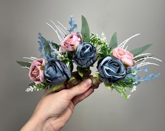 May include: A floral crown featuring blue and pink roses, green leaves, and small white and blue accents. The flowers are arranged on a brown base, creating a delicate and romantic headpiece.