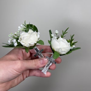 Könnte beinhalten: Zwei weiße Blumen-Ansteckblumen mit grünen Blättern und kleinen weißen Blüten. Jede Ansteckblume hat einen silbernen Kreuz-Anhänger.