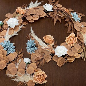 May include: A decorative wreath featuring brown eucalyptus leaves, white, peach, and blue flowers, and feathery accents. The wreath is arranged in a circular shape, perfect for home decor.
