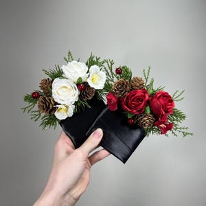 Puede incluir: Dos arreglos florales de satén negro, cada uno adornado con rosas blancas y rojas, piñas y vegetación. Los arreglos se sostienen en una mano, mostrando el contraste entre la tela oscura y las flores vibrantes.