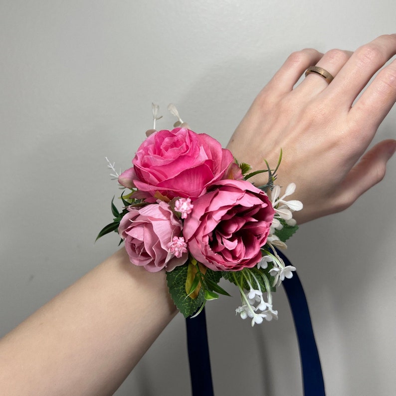 May include: A pink and white floral wrist corsage with a navy blue ribbon. The corsage features three pink roses and white baby's breath.