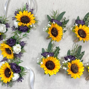 May include: An assortment of sunflower and floral boutonnieres and wrist corsages. The boutonnieres feature yellow sunflowers with dark brown centers, purple flowers, white flowers, and green foliage. The wrist corsages have white ribbons.