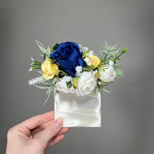 Puede incluir: Un pequeño ramo de flores artificiales azules y amarillas dispuestas en una bolsa de satén blanco.