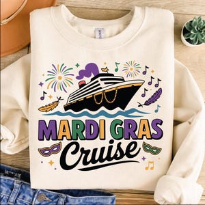May include: Cream-colored sweatshirt with a Mardi Gras Cruise design. The graphic features a black cruise ship, musical notes, fireworks, and the words "Mardi Gras Cruise" in purple, green, and black. Decorative masks and feathers complete the design.