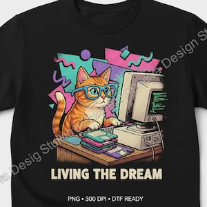 May include: Black t-shirt featuring a cartoon cat wearing glasses and working on a computer. The cat is orange and white, and the design includes the text "LIVING THE DREAM" in white letters. The design has a retro 80s or 90s vibe.