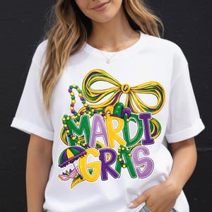 May include: White t-shirt with a colorful Mardi Gras graphic. The design features the words "Mardi Gras" in green, yellow, and purple, along with a bow, beads, and a mask.