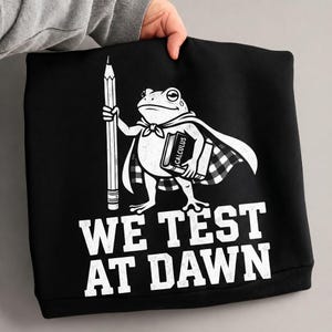 May include: Black sweatshirt featuring a white cartoon frog wearing a cape and holding a pencil and a book labeled "Calculus." The text "WE TEST AT DAWN" is printed below the frog in white block letters.