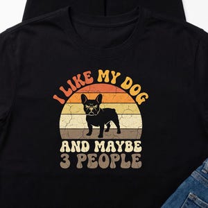 May include: Black t-shirt with a retro design featuring a French Bulldog silhouette inside a sunset graphic. Text reads "I LIKE MY DOG AND MAYBE 3 PEOPLE" in a vintage font. A casual, dog-themed apparel item.