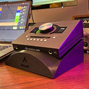 May include: A Universal Audio audio interface with a large silver knob and a purple stand. The interface is connected to a laptop computer with a music production program open on the screen. The interface is also connected to a black speaker.