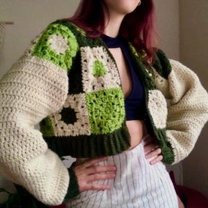 Könnte beinhalten: Eine gehäkelte Strickjacke in Grün, Weiß und Creme mit Patchwork-Muster. Die Strickjacke ist kurz und hat lange Ärmel.