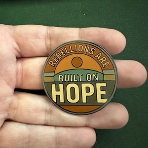 May include: A round, bronze-colored coin with the words "REBELLIONS ARE BUILT ON HOPE" in a circular design. The coin features a stylized landscape with a sun and earth tones.