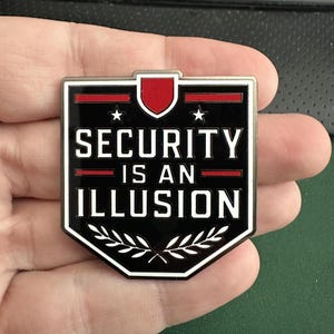 Op de afbeelding: Een zwart-witte schildvormige pin met de tekst "SECURITY IS AN ILLUSION". De pin heeft een rode accentkleur aan de bovenkant en een laurierkransontwerp aan de onderkant. Twee sterren bevinden zich in de bovenhoeken.