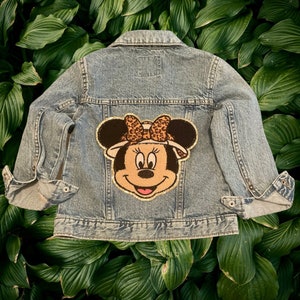 May include: A blue denim jacket with a brown and gold Minnie Mouse patch on the back. The patch features Minnie Mouse wearing a safari hat and a leopard print bow.