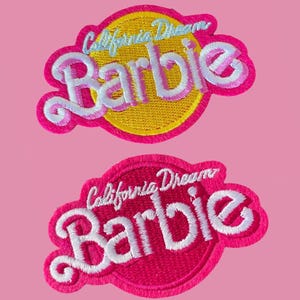 May include: Two embroidered patches with the word "Barbie" in a cursive font. One patch has a yellow circle with the text "California Dream" in light blue. The other patch is a pink circle with white text.