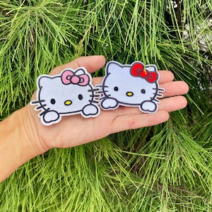 K&ouml;nnte beinhalten: Zwei wei&szlig;e, gestickte B&uuml;gelbilder von Hello Kitty, eines mit einer rosa Schleife und das andere mit einer roten Schleife.