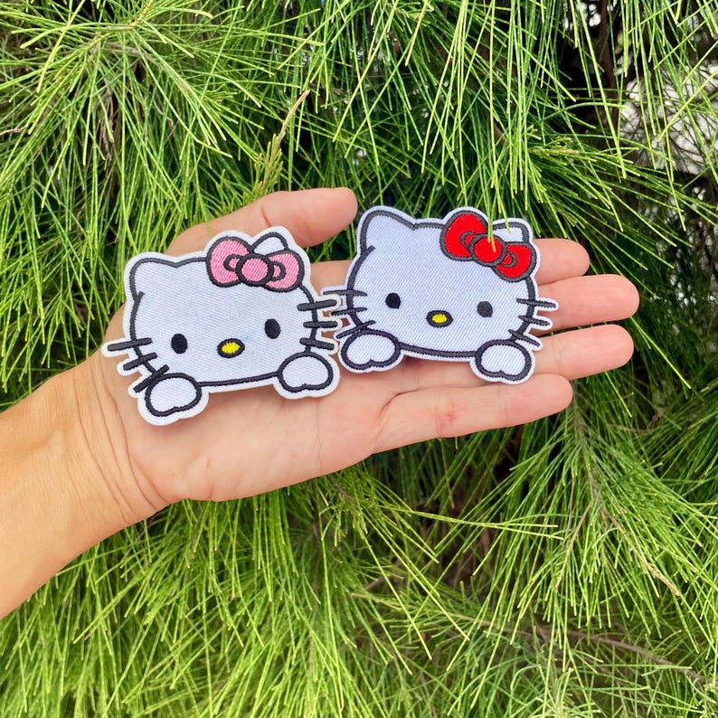 K&ouml;nnte beinhalten: Zwei wei&szlig;e, gestickte B&uuml;gelbilder von Hello Kitty, eines mit einer rosa Schleife und das andere mit einer roten Schleife.