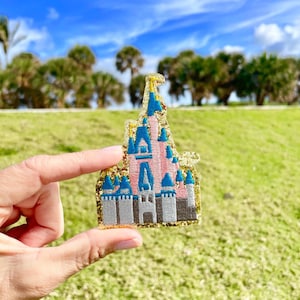 May include: An embroidered patch shaped like a castle with a pink facade, blue roofs, and a grey base. The entire design is outlined and accented with a shimmering gold glitter border, creating a distinct silhouette against the background.